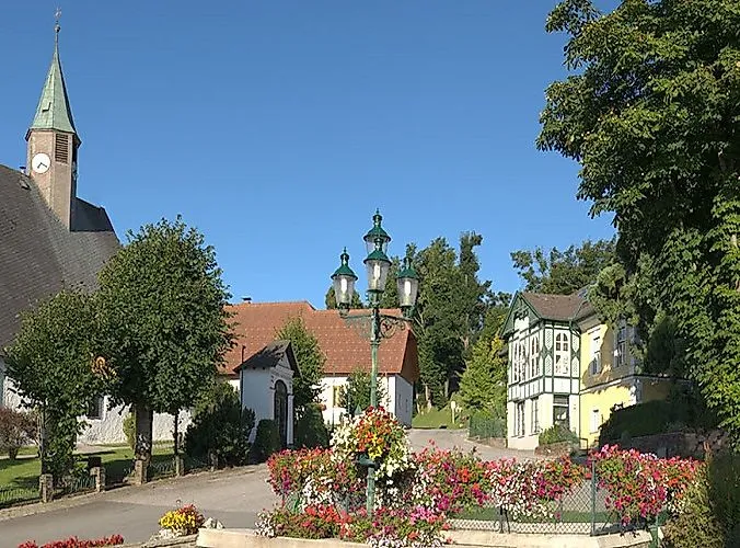 Sommerlicher Dorfplatz in Mönichkirchen mit Blumenbeeten, historischen Gebäuden und blauem Himmel.
