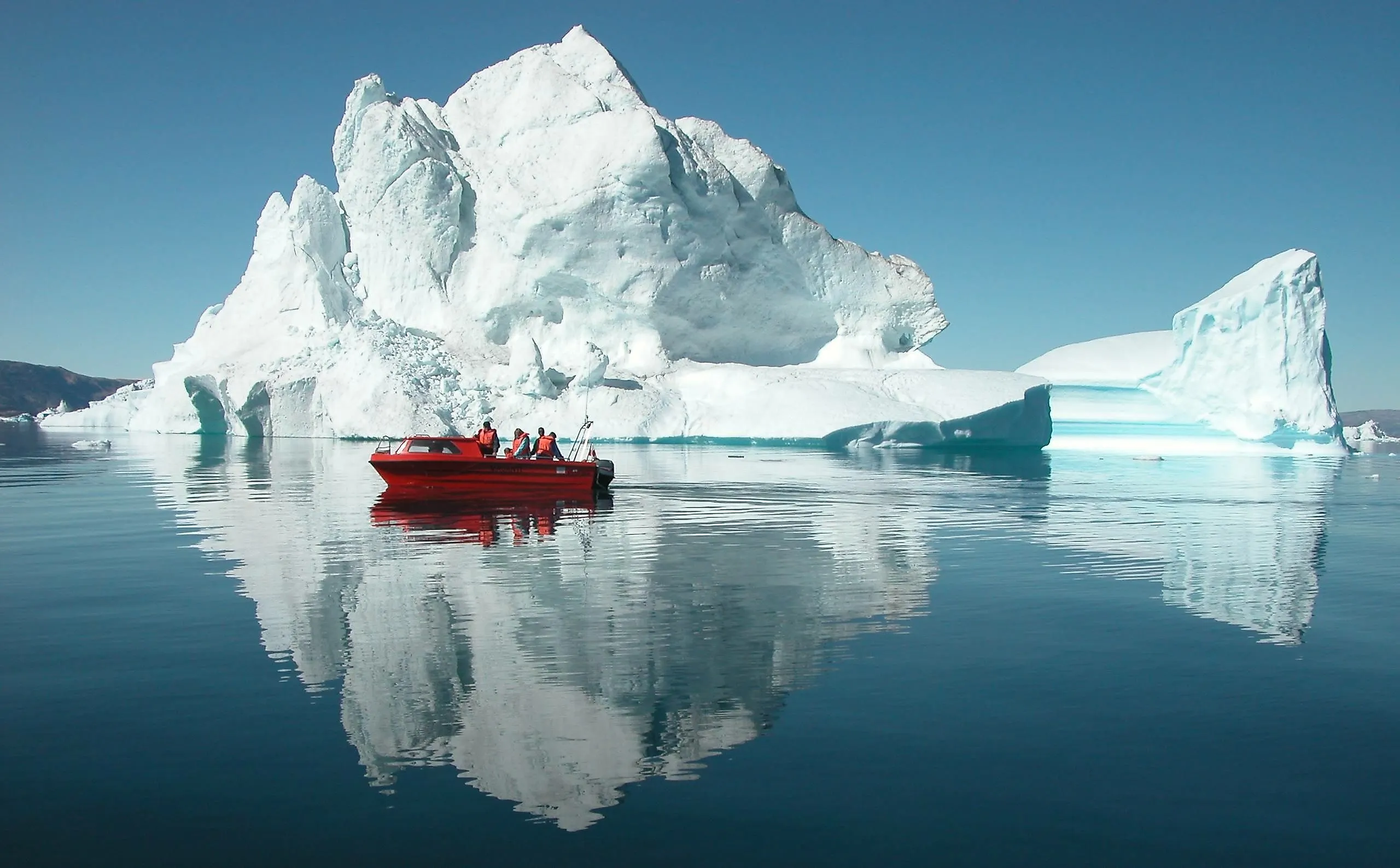 Personen im roten Boot vor einem riesigen Eisberg in Ostgrönland mit klarer Spiegelung