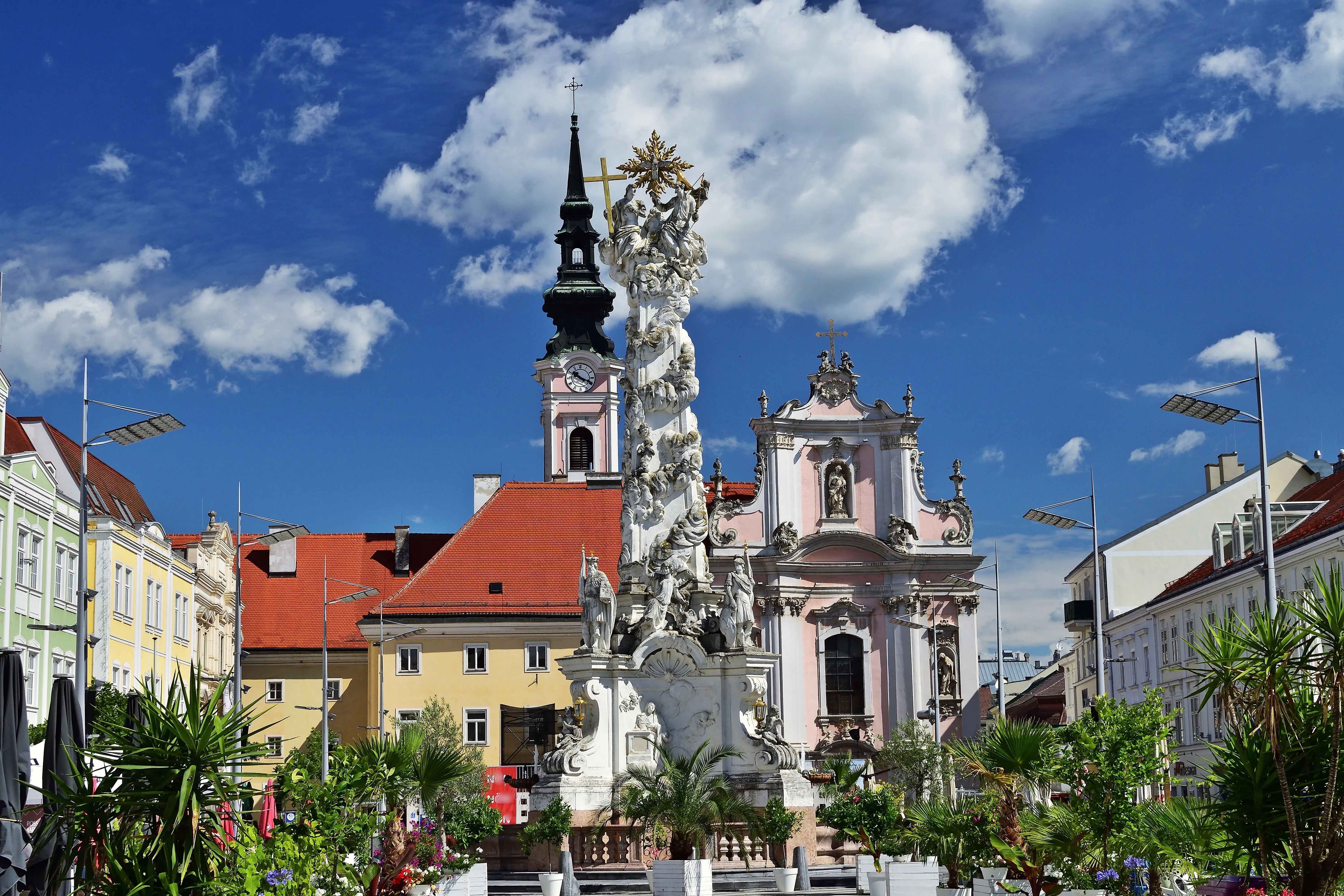 Colonne historique de la Trinité devant l'église baroque des Franciscains au centre de St. Pölten, entourée de maisons bourgeoises colorées et de décorations florales estivales.