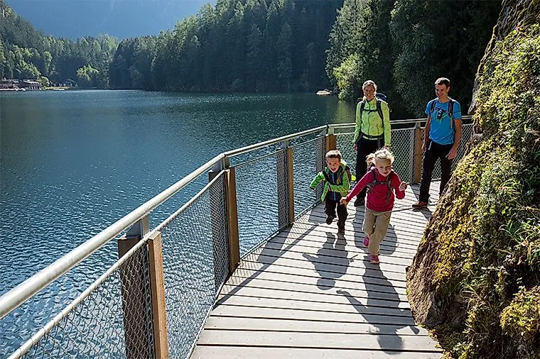 Familie wandert auf einem Holzsteg entlang des Piburger Sees in Tirol mit Blick auf Wasser und Waldkulisse.