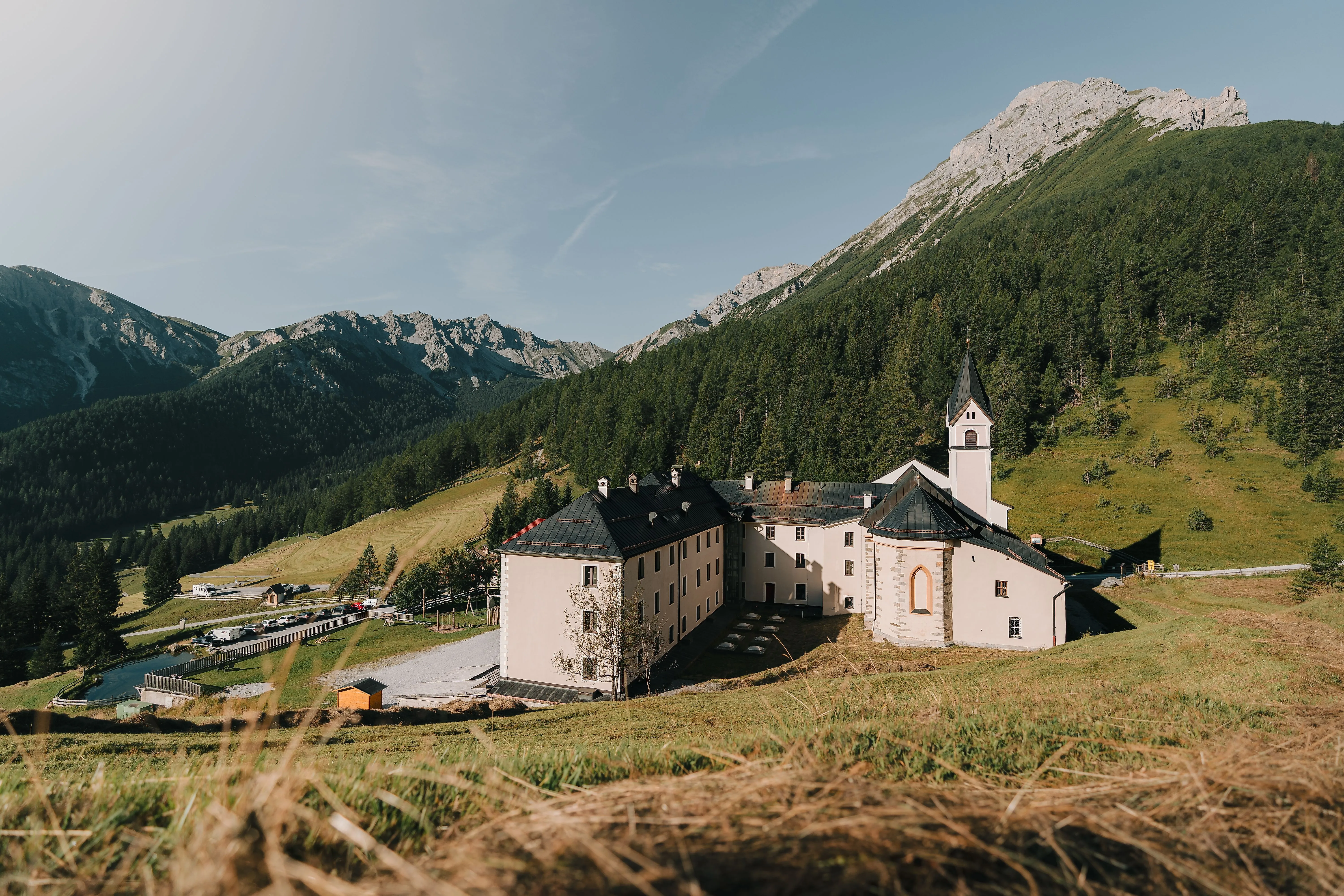 Das Kloster Maria Waldrast inmitten grüner Almwiesen vor imposanter Bergkulisse an einem sonnigen Sommertag.