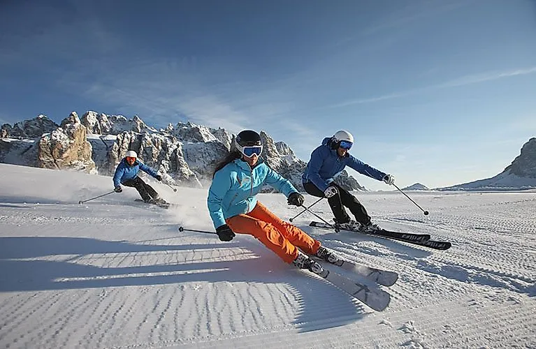 Drei Skifahrer carven über die Piste vor verschneiten Gipfeln der Dolomiten