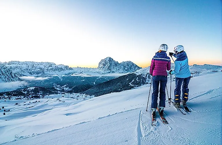 Zwei Skifahrer:innen auf verschneiter Piste mit Blick auf die Dolomiten bei Sonnenaufgang