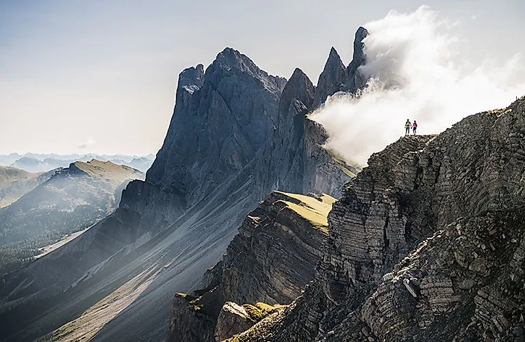 Zwei Wanderer:innen auf einem steilen Dolomitenkamm umgeben von Wolken und Felsen