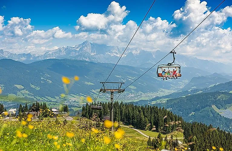 Letní horská krajina ve Flachau s rozkvetlými loukami, lanovkou a výhledem na Alpy v pozadí.