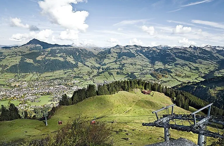 Weitläufiger Blick auf die grünen Kitzbüheler Alpen mit Wiesen, kleinen Wäldern und verstreuten Dörfern; im Vordergrund eine Seilbahn mit roten Gondeln.